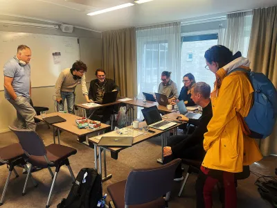 A group of nine people gather in a classroom around tables with laptops, engaged in discussion near a whiteboard and windows