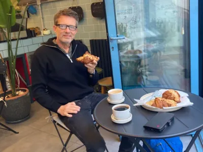 Man in glasses eating a pastry at a small table in a café, with coffee cups and a plate of pastries nearby