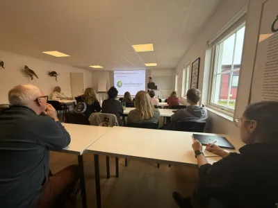 A classroom with seated attendees facing a presenter at a screen displaying "biodiverso+" content near windows overlooking a…